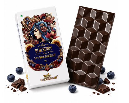 A bar of chocolate and its packaging box on a white background, styled with whole and broken blueberries and chocolate crumbs. The box is white with a colorful floral illustration and reads 'Blueberry 5th Dark Chocolate'.