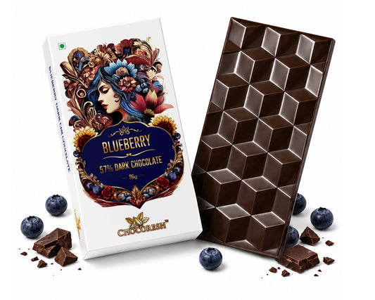 A bar of chocolate and its packaging box on a white background, styled with whole and broken blueberries and chocolate crumbs. The box is white with a colorful floral illustration and reads 'Blueberry 5th Dark Chocolate'.
